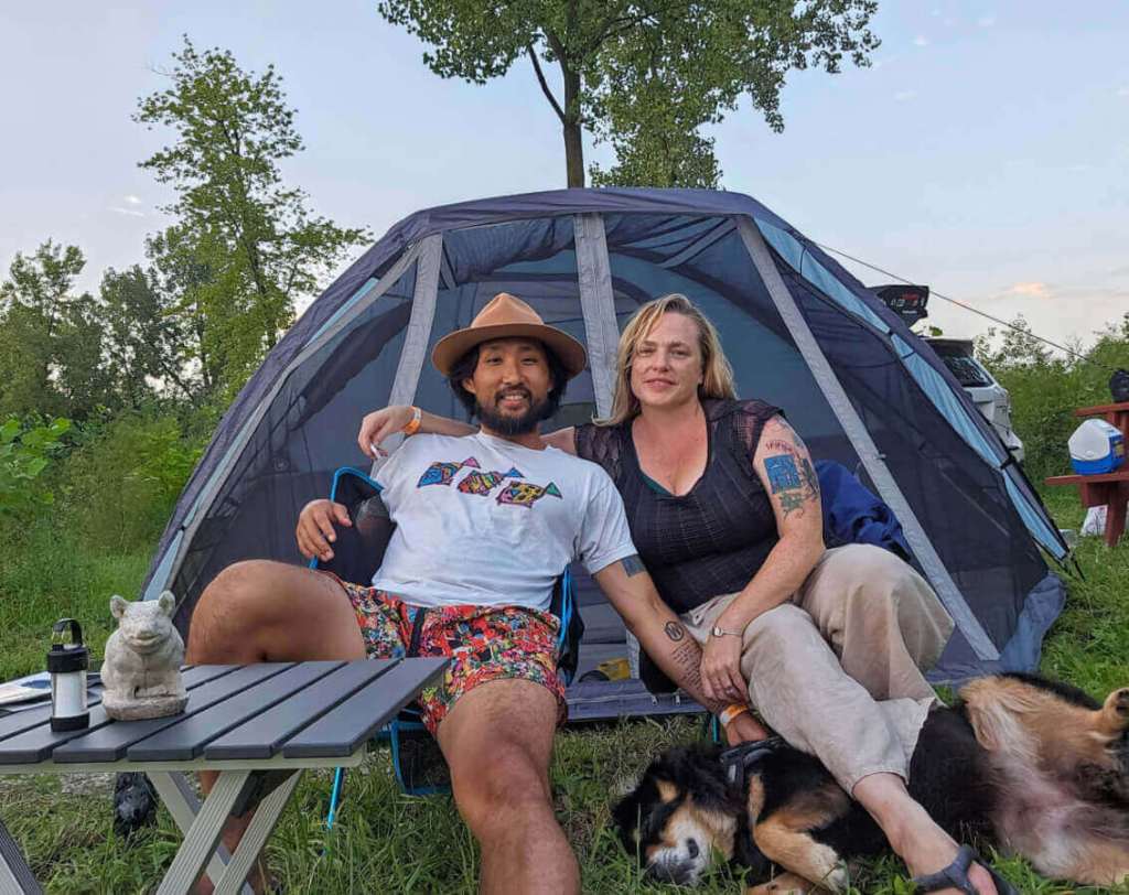 A bearded Korean man in a tan hat, a white woman with tattoos and their dog posing in front of a tent.