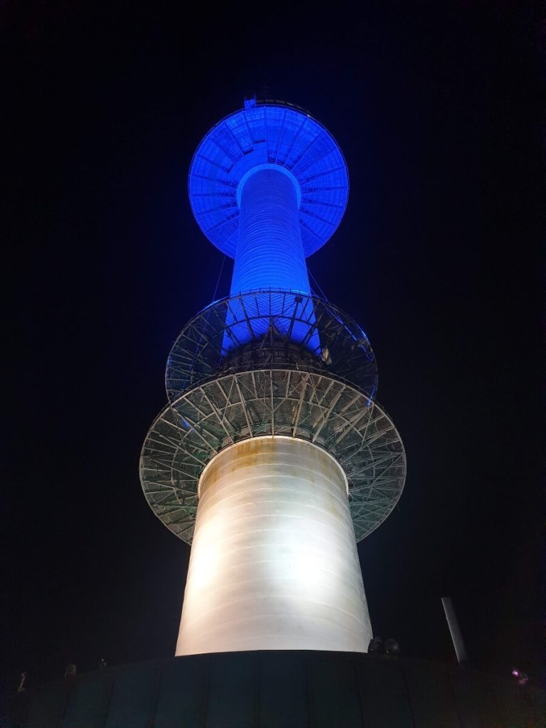 A photograph of the Seoul Tower at night lit up with blue light. Taken at Namsan Park in Seoul, South Korea.