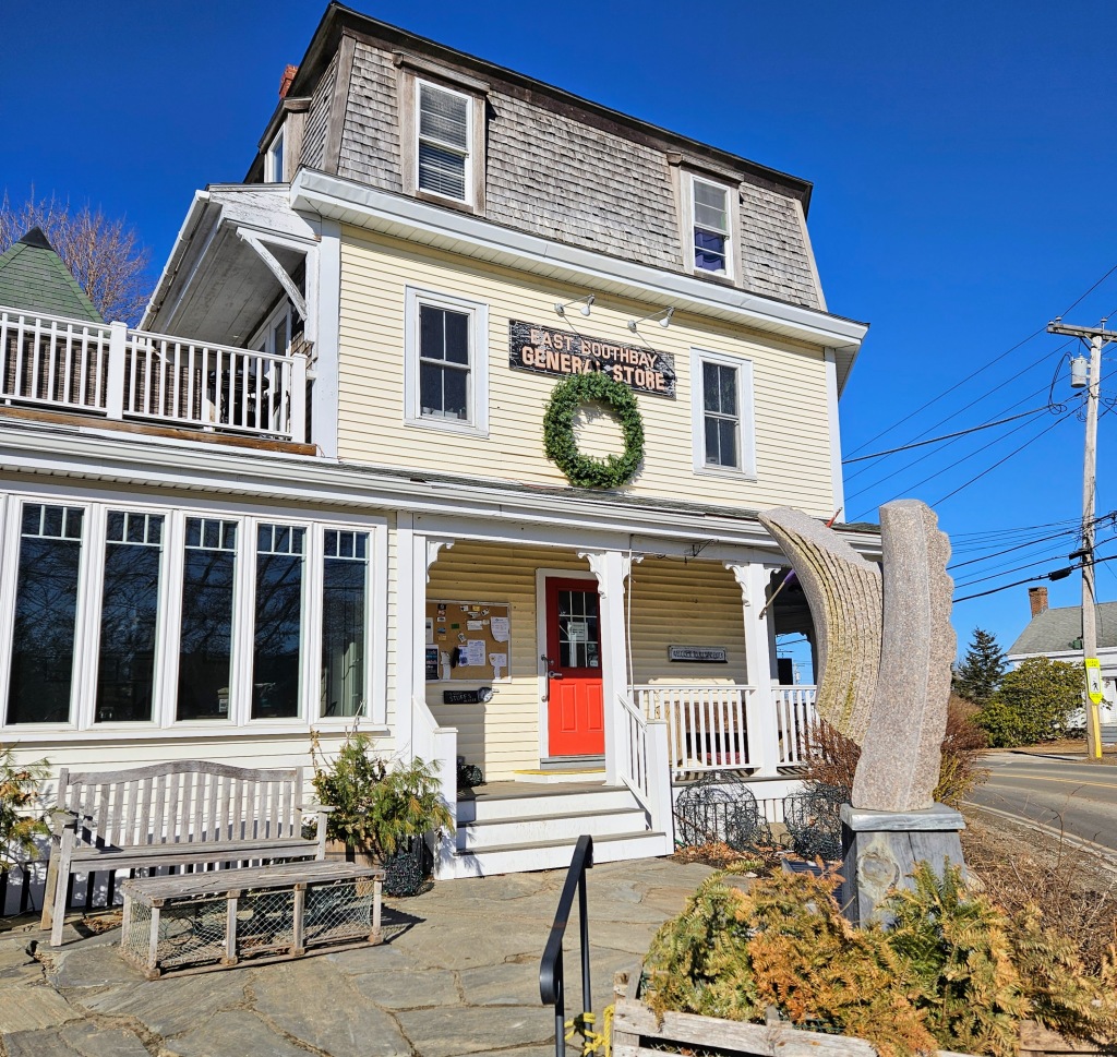 A house that is built in the Queen Anne style with a mansard roof. The house is a lemon yellow and has a red front door. The sign on the house says, East Boothbay General Store. Under the sign is a giant wreath. In front of the house is a stone sculpture.